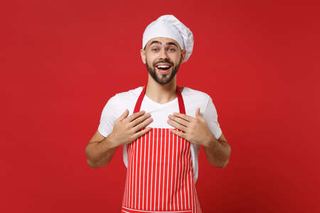 Excited young bearded male chef cook or baker man in striped apron white t-shirt toque chefs hat posing isolated on red background. Cooking food concept. Mock up copy space. Keeping hands on chest.の写真素材