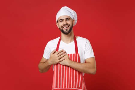 Smiling young bearded male chef cook or baker man in striped apron white t-shirt toque chefs hat posing isolated on red background. Cooking food concept. Mock up copy space. Holding hands on heart.の写真素材