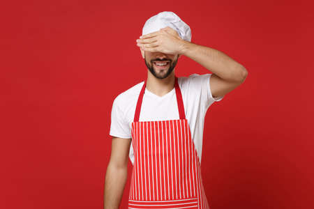 Smiling young bearded male chef cook or baker man in striped apron white t-shirt toque chefs hat posing isolated on red background. Cooking food concept. Mock up copy space. Covering eyes with palm.の写真素材