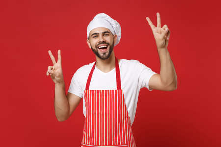 Cheerful young bearded male chef cook or baker man in striped apron white t-shirt toque chefs hat posing isolated on red background. Cooking food concept. Mock up copy space. Showing victory sign.の写真素材