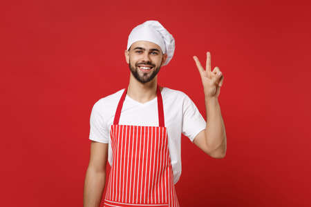 Smiling young bearded male chef cook or baker man in striped apron white t-shirt toque chefs hat posing isolated on red wall background. Cooking food concept. Mock up copy space. Showing victory sign.の写真素材