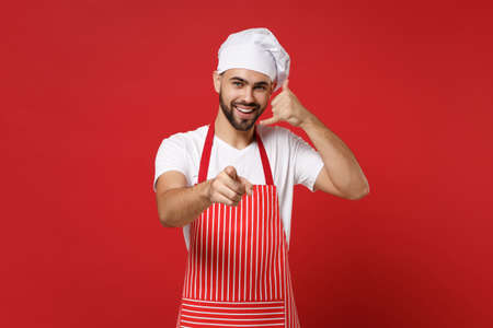 Bearded male chef cook or baker man in striped apron toque chefs hat posing isolated on red background. Cooking food concept. Doing phone gesture like says call me back, pointing finger on camera.の写真素材
