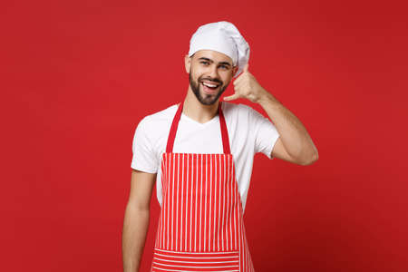 Young bearded male chef cook or baker man in striped apron toque chefs hat posing isolated on red wall background. Cooking food concept. Mock up copy space. Doing phone gesture like says call me back.の写真素材