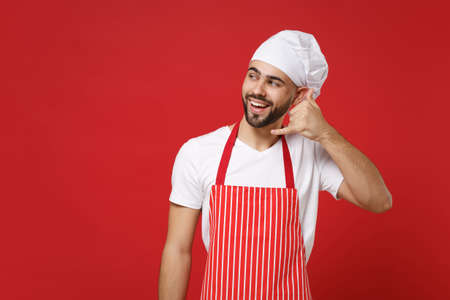 Young bearded male chef cook or baker man in striped apron toque chefs hat posing isolated on red wall background. Cooking food concept. Mock up copy space. Doing phone gesture like says call me back.の写真素材