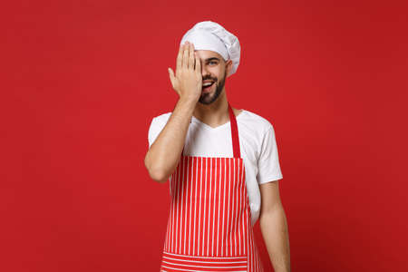 Cheerful young bearded male chef cook or baker man in striped apron white t-shirt toque chefs hat posing isolated on red background. Cooking food concept. Mock up copy space. Covering eyes with palm.の写真素材