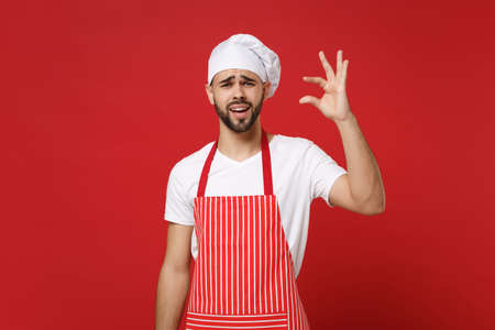 Young bearded male chef cook or baker man in striped apron toque chefs hat posing isolated on red background. Cooking food concept. Mock up copy space. Gesturing demonstrating size with workspace.の写真素材