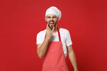 Young bearded male chef cook or baker man in striped apron toque chefs hat posing isolated on red background. Cooking food concept. Mock up copy space. Showing tongue, depicting heavy metal rock sign.の写真素材