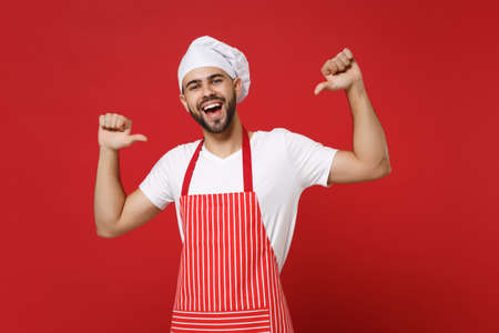 Cheerful young bearded male chef cook or baker man in striped apron white t-shirt toque chefs hat posing isolated on red background. Cooking food concept. Mock up copy space. Point thumbs on himself.の写真素材
