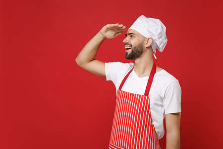 Bearded male chef cook or baker man in striped apron toque chefs hat posing isolated on red background. Cooking food concept. Mock up copy space. Holding hand at forehead looking far away distance.の写真素材