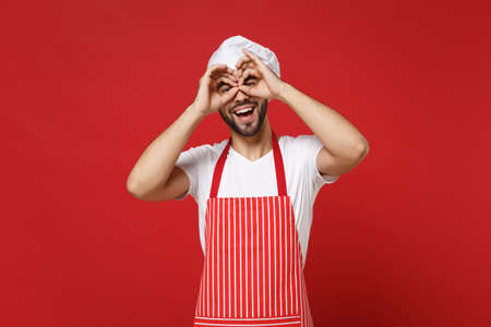 Bearded male chef cook or baker man in striped apron toque chefs hat posing isolated on red background. Cooking food concept. Mock up copy space. Hold hands near eyes, imitating glasses or binoculars.の写真素材
