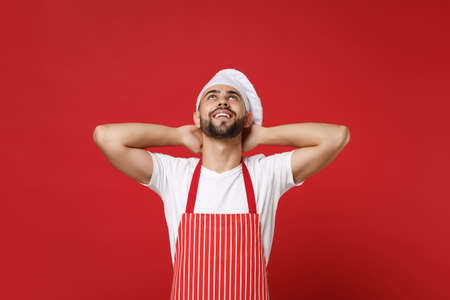 Young bearded male chef cook or baker man in striped apron white t-shirt toque chefs hat posing isolated on red background. Cooking food concept. Mock up copy space. Looking up with hands behind head.の写真素材