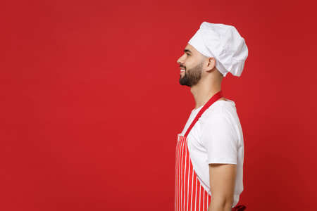 Side view of smiling young bearded male chef cook or baker man in striped apron white t-shirt toque chefs hat posing isolated on red background in studio. Cooking food concept. Mock up copy space.の写真素材