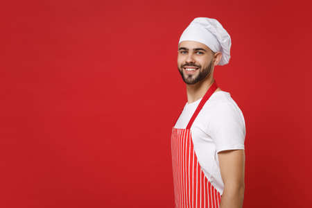Side view of young bearded male chef cook or baker man in striped apron white t-shirt toque chefs hat posing isolated on red wall background. Cooking food concept. Mock up copy space. Looking camera.の写真素材