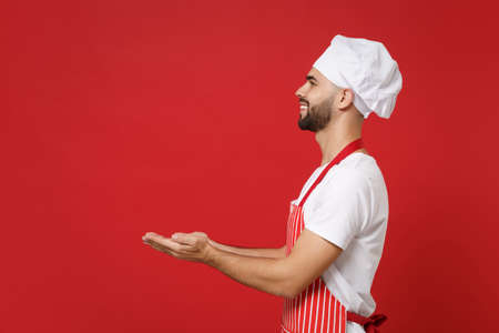 Side view of bearded male chef cook or baker man in striped apron white t-shirt toque chefs hat posing isolated on red background. Cooking food concept. Mock up copy space. Holding something in hands.の写真素材