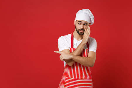 Bearded male chef cook or baker man in striped apron toque chefs hat posing isolated on red background. Cooking food concept. Whispers gossip tell secret with hand gesture pointing index finger aside.の写真素材