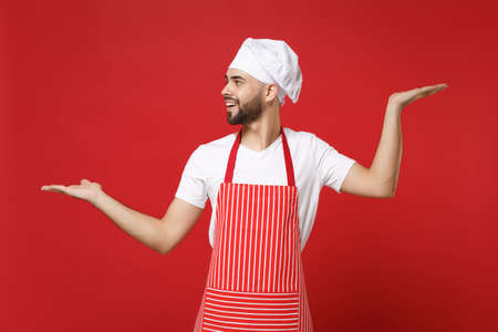 Young bearded male chef cook or baker man in striped apron toque chefs hat posing isolated on red background. Cooking food concept. Mock up copy space. Spreading, pointing hands aside, hold something.の写真素材