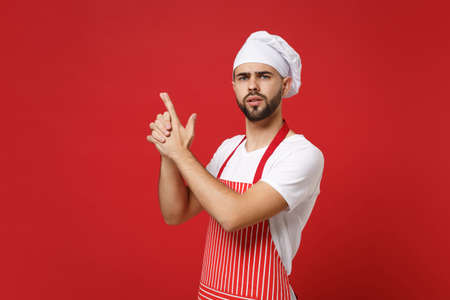 Concerned young bearded male chef cook or baker man in striped apron white t-shirt toque chefs hat posing isolated on red background. Cooking food concept. Mock up copy space. Holding hands like gun.の写真素材