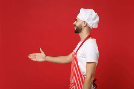 Side view of young male chef cook or baker man in striped apron toque chefs hat posing isolated on red background. Cooking food concept. Mock up copy space. Stand with outstretched hand for greeting.の写真素材