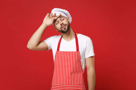 Tired exhausted young male chef cook or baker man in striped apron toque chefs hat posing isolated on red background. Cooking food concept. Mock up copy space. Put hand on head, keeping eyes closed.の写真素材