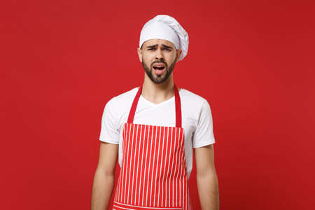 Perplexed confused young bearded male chef cook or baker man in striped apron white t-shirt toque chefs hat posing isolated on red background studio portrait. Cooking food concept. Mock up copy space.の写真素材