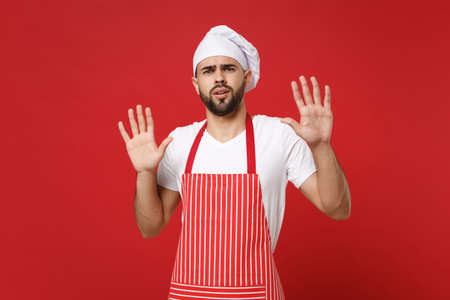 Shocked scared bearded male chef cook or baker man in striped apron t-shirt toque chefs hat posing isolated on red background. Cooking food concept. Mock up copy space. Rising hands, showing palms.の写真素材