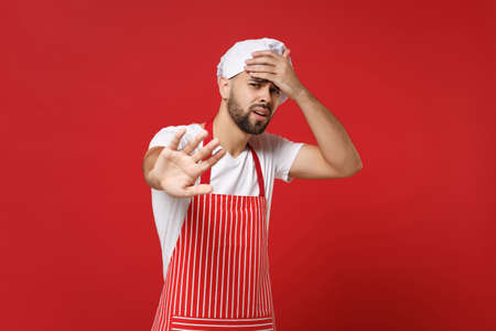 Young bearded male chef cook or baker man in striped apron toque chefs hat posing isolated on red background. Cooking food concept. Mock up copy space. Put hand on head showing stop gesture with palm.の写真素材