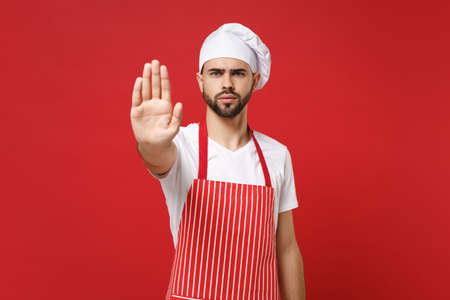 Serious bearded male chef cook or baker man in striped apron white t-shirt toque chefs hat posing isolated on red background. Cooking food concept. Mock up copy space. Showing stop gesture with palm.の写真素材