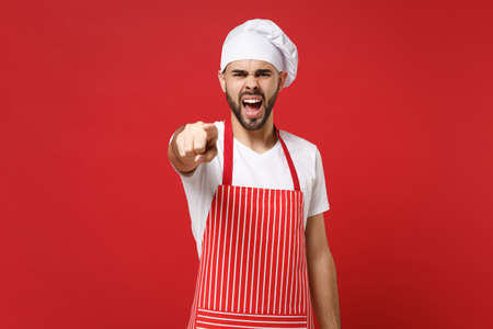 Angry bearded male chef cook or baker man in striped apron toque chefs hat posing isolated on red background. Cooking food concept. Mock up copy space. Screaming swearing point index finger on camera.の写真素材