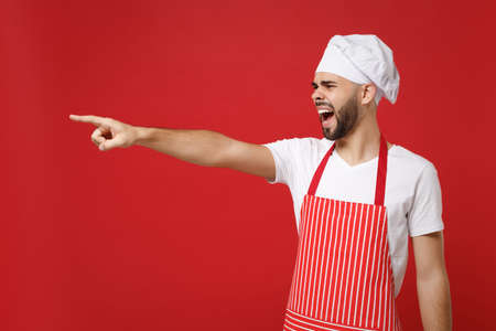 Angry bearded male chef cook or baker man in striped apron toque chefs hat posing isolated on red background. Cooking food concept. Mock up copy space. Screaming swearing pointing index finger aside.の写真素材