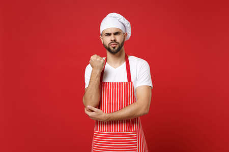 Angry young bearded male chef cook or baker man in striped apron white t-shirt toque chefs hat posing isolated on red background in studio. Cooking food concept. Mock up copy space. Clenching fist.の写真素材
