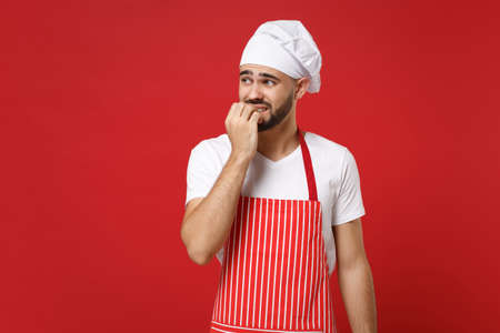 Worried young bearded male chef cook or baker man in striped apron white t-shirt toque chefs hat posing isolated on red background in studio. Cooking food concept. Mock up copy space. Gnawing nails.の写真素材