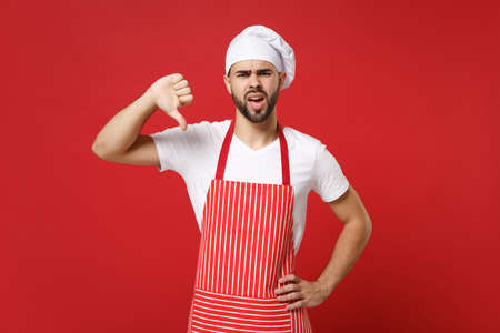 Displeased young bearded male chef cook or baker man in striped apron white t-shirt toque chefs hat posing isolated on red background. Cooking food concept. Mock up copy space. Showing thumb down.の写真素材