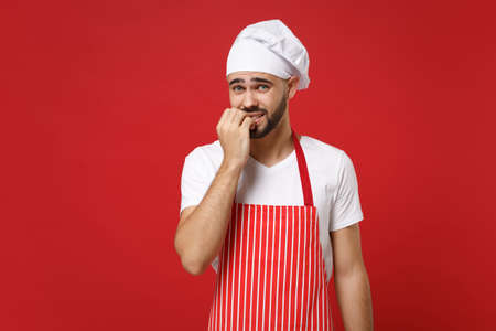 Nervous young bearded male chef cook or baker man in striped apron white t-shirt toque chefs hat posing isolated on red background in studio. Cooking food concept. Mock up copy space. Gnawing nails.の写真素材