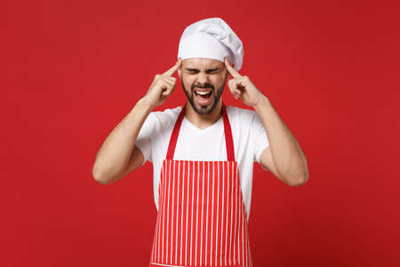 Frustrated male chef cook or baker man in striped apron toque chefs hat posing isolated on red background. Cooking food concept. Mock up copy space. Screaming, keeping eyes closed, put hands on head.の写真素材