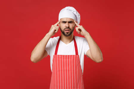Preoccupied young bearded male chef cook or baker man in striped apron toque chefs hat posing isolated on red wall background. Cooking food concept. Mock up copy space. Pointing index fingers on head.の写真素材