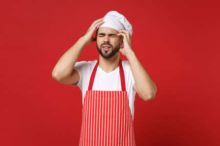 Young bearded male chef cook or baker man in striped apron toque chefs hat posing isolated on red wall background. Cooking food concept. Mock up copy space. Pointing hands on head keeping eyes closed.の写真素材