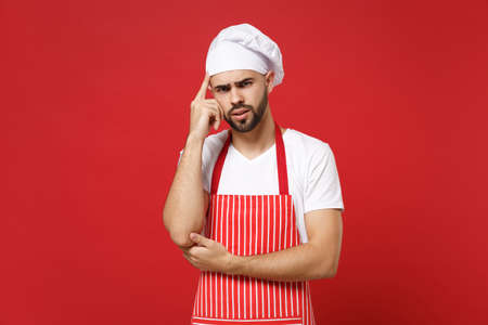 Pensive young bearded male chef cook or baker man in striped apron toque chefs hat posing isolated on red background in studio. Cooking food concept. Mock up copy space. Pointing index finger on head.の写真素材