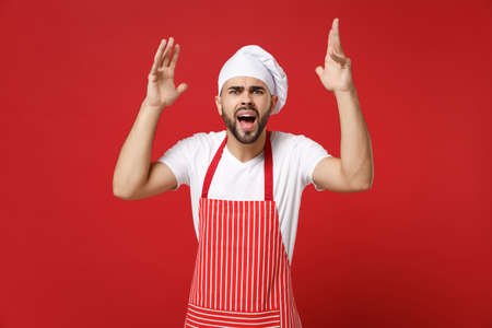 Irritated young bearded male chef cook or baker man in striped apron toque chefs hat posing isolated on red background. Cooking food concept. Mock up copy space. Screaming, swearing, spreading hands.の写真素材