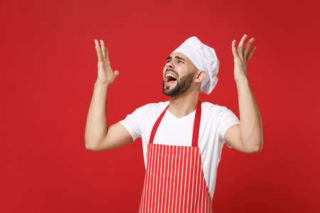 Angry young bearded male chef cook or baker man in striped apron toque chefs hat posing isolated on red wall background. Cooking food concept. Mock up copy space. Screaming, swearing, spreading hands.の写真素材