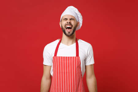 Frustrated young bearded male chef cook or baker man in striped apron toque chefs hat posing isolated on red wall background. Cooking food concept. Mock up copy space. Screaming, keeping eyes closed.の写真素材