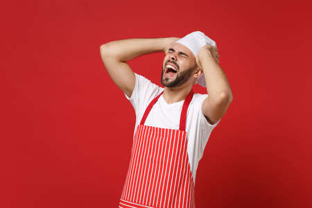 Frustrated male chef cook or baker man in striped apron toque chefs hat posing isolated on red background. Cooking food concept. Mock up copy space. Screaming, keeping eyes closed, put hands on head.の写真素材
