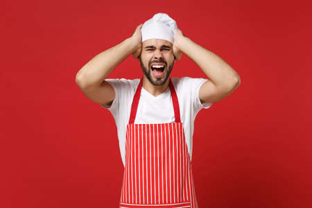 Crazy young bearded male chef cook or baker man in striped apron white t-shirt toque chefs hat posing isolated on red background. Cooking food concept. Mock up copy space. Screaming put hands on head.の写真素材