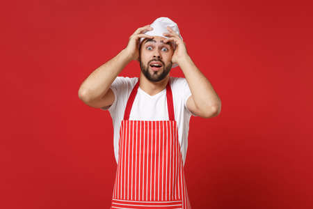 Shocked young bearded male chef cook or baker man in striped apron white t-shirt toque chefs hat posing isolated on red background. Cooking food concept. Mock up copy space. Putting hands on head.の写真素材