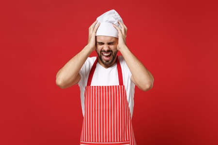 Bearded male chef cook or baker man in striped apron toque chefs hat posing isolated on red background. Cooking food concept. Mock up copy space. Screaming, keeping eyes closed, putting hands on head.の写真素材