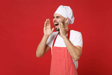 Angry young bearded male chef cook or baker man in striped apron toque chefs hat posing isolated on red background. Cooking food concept. Mock up copy space. Screaming with hand gesture looking aside.の写真素材