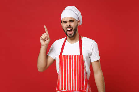 Angry bearded male chef cook or baker man in striped apron toque chefs hat posing isolated on red background. Cooking food concept. Mock up copy space. Screaming, swearing, pointing index finger up.の写真素材