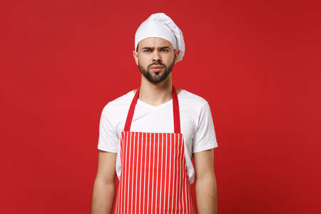 Puzzled perplexed young bearded male chef cook or baker man in striped apron white t-shirt toque chefs hat posing isolated on red background, studio portrait. Cooking food concept. Mock up copy space.の写真素材