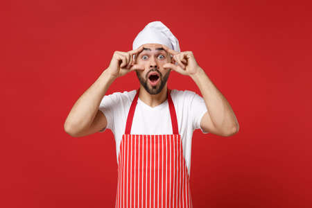 Shocked young bearded male chef cook or baker man in striped apron toque chefs hat posing isolated on red background. Cooking food concept. Mock up copy space. Keeping mouth open, stretching eyelids.の写真素材