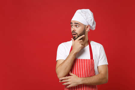 Shocked young bearded male chef cook or baker man in striped apron toque chefs hat posing isolated on red background. Cooking food concept. Mock up copy space. Put hand prop up on chin, looking aside.の写真素材