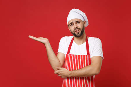 Concerned young bearded male chef cook or baker man in striped apron white t-shirt toque chefs hat posing isolated on red background. Cooking food concept. Mock up copy space. Pointing hand aside.の写真素材